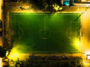 Aerial night shot of a fully lit soccer field, surrounded by trees and small buildings. World Cup rivalries